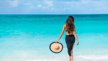 Woman from behind on a white-sand beach in a black sarong cover-up holding a straw sun hat, turquoise sea in front — Donjane beach cover-ups for women.